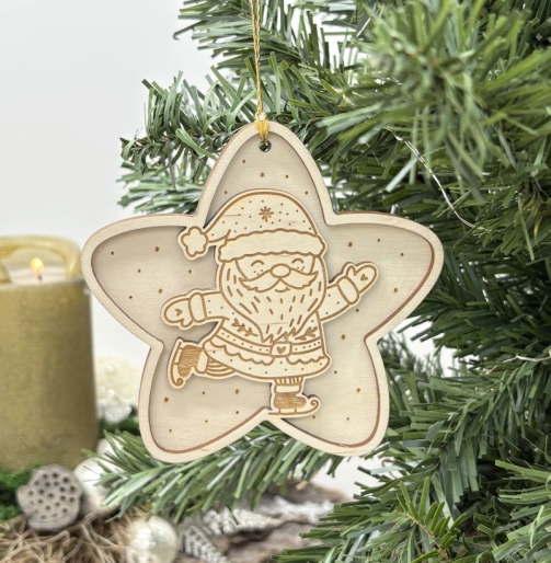 Frontal fotografierter Weihnachtsanhänger aus hellem Holz in Sternform mit graviertem Weihnachtsmann auf Schlittschuhen, Weihnachtsmütze, Bart und kleinen Sternen, aufgehängt zwischen grünen Tannenzweigen.